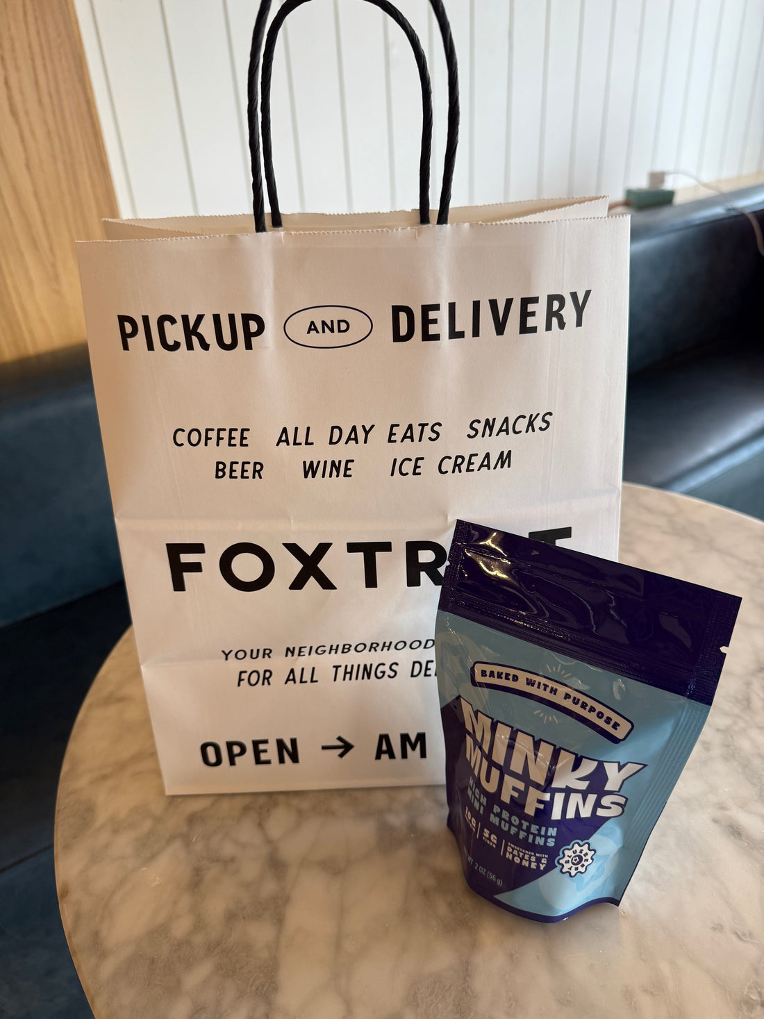A Foxtrot togo bag with a bag of Minky Muffins high protein blueberry mini muffins sitting on a table.