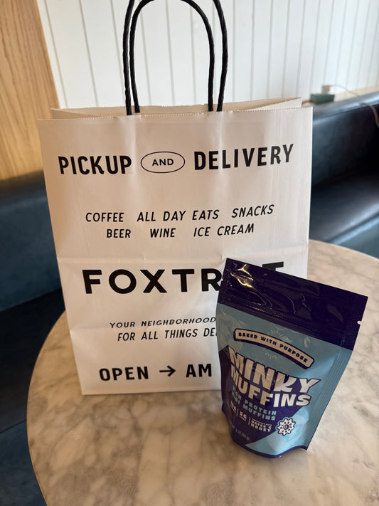 A Foxtrot togo bag with a bag of Minky Muffins high protein blueberry mini muffins sitting on a table.