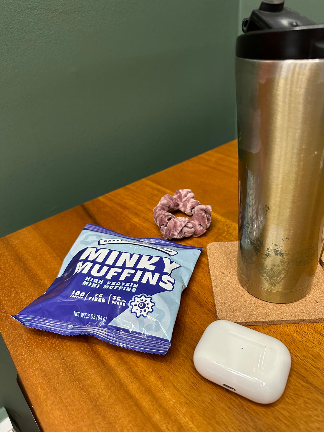 A bag of mini muffins on a desk next to a hair scrunchy, an Airpods case, and a thermos for coffee. 